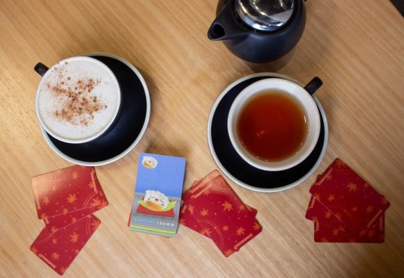 A table with a card game scattered around a mug full of tea, and mug of coffee.