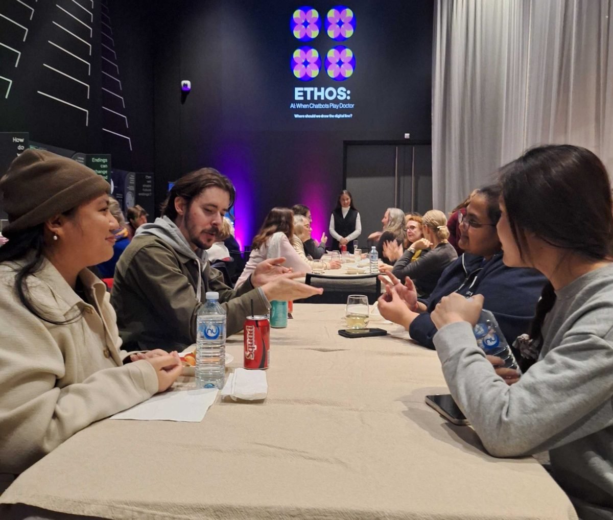 Four participants sitting at a trestle table with drinks in a brightly lit room discussing the ethics of using AI chatbots for health advice. In the background there are more tables with participants.