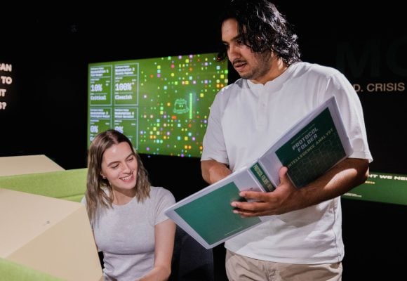 Person is standing up holding a folder. Another person is sitting at a computer looking over to the folder.