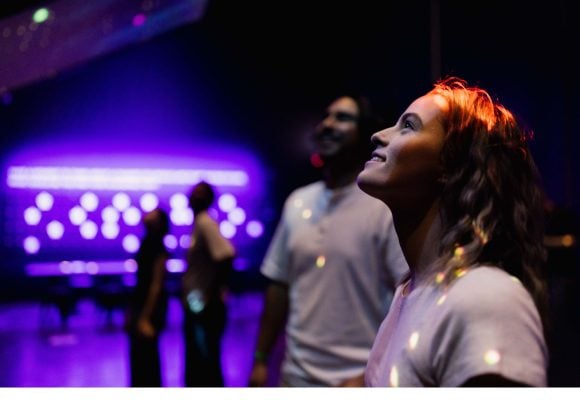 A person is looking up, with stars projected on to their face. The background is blurry with wall lit up purple.
