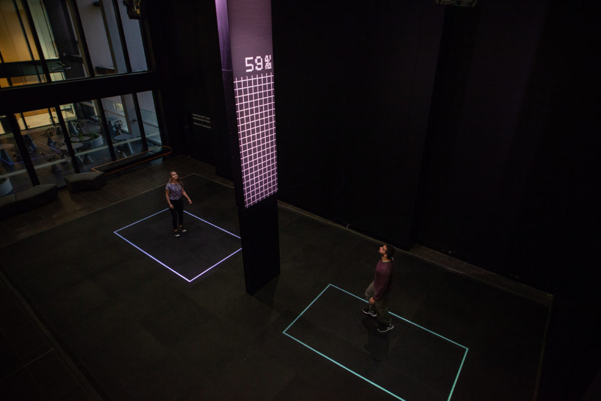 Two people stand in a large room. There is rubber gym matting on the floor and a tall black tower between them. The tower has many white dots that have flickered to display a task bar filled to 59%.