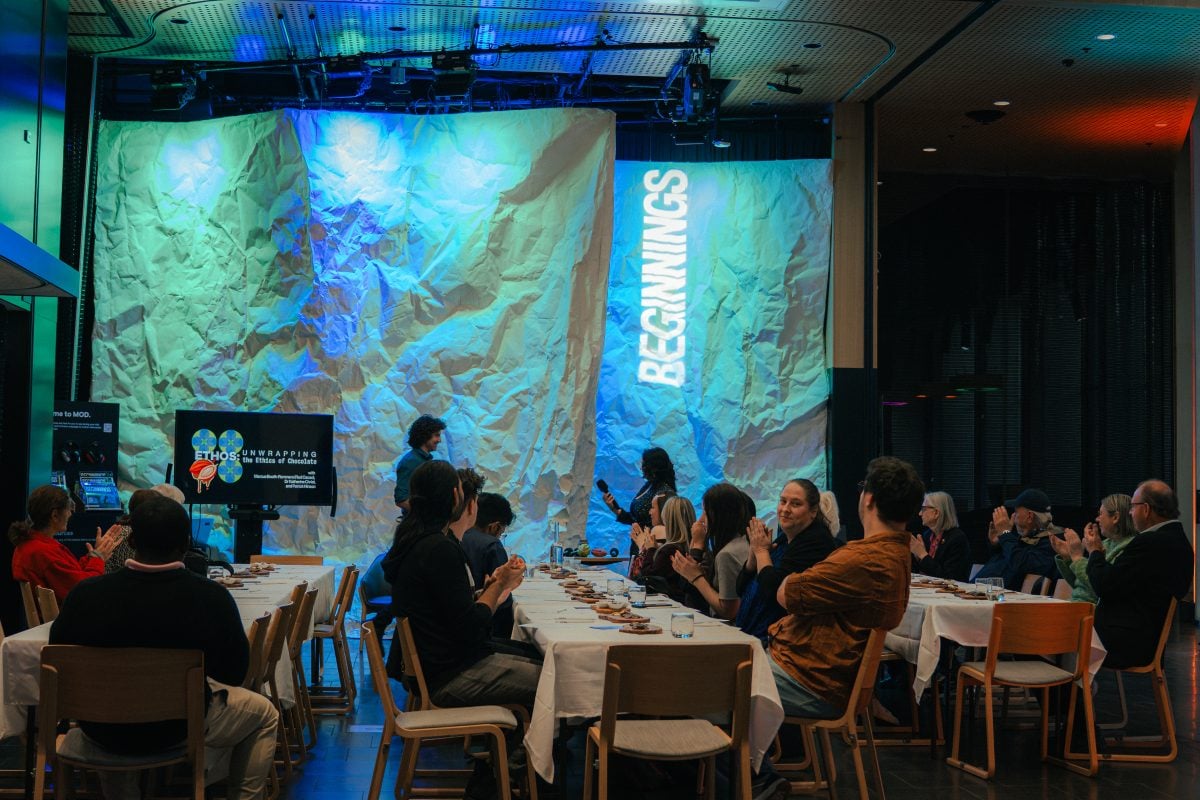 There are three long tables with white table cloths with people sitting around them. In the background there is the entrance to MOD.'s BEGINNINGS exhibition, lit with blue and green light.