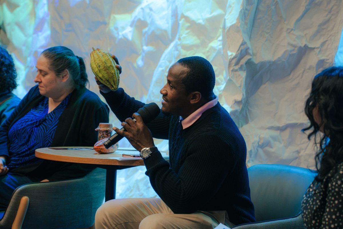 There is a person speaking on a panel, they are holding a cocoa pod with one hand and speaking into a microphone with the other.