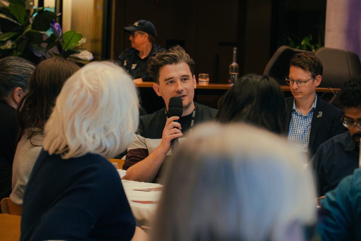 A person sitting at a table amongst a group of others is holding a microphone and speaking.