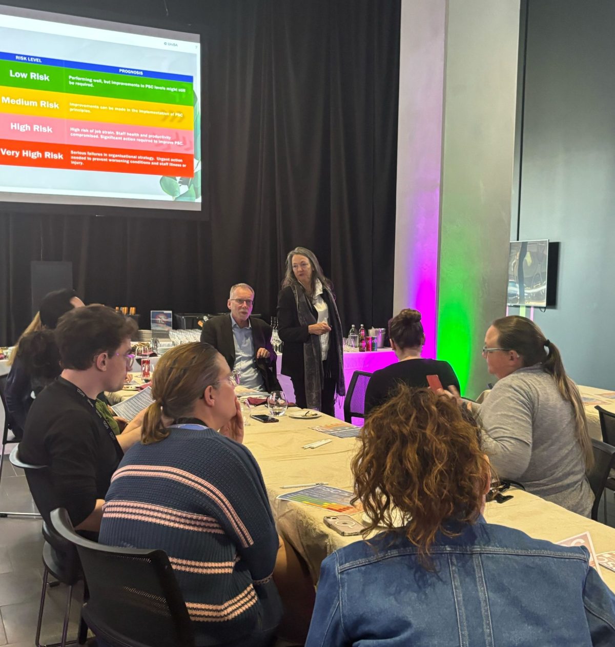 Ten forum participants and panelists in a brightly lit room sitting around trestle tables. They have pencils and a PSC questionnaire on A4 paper in front of them.