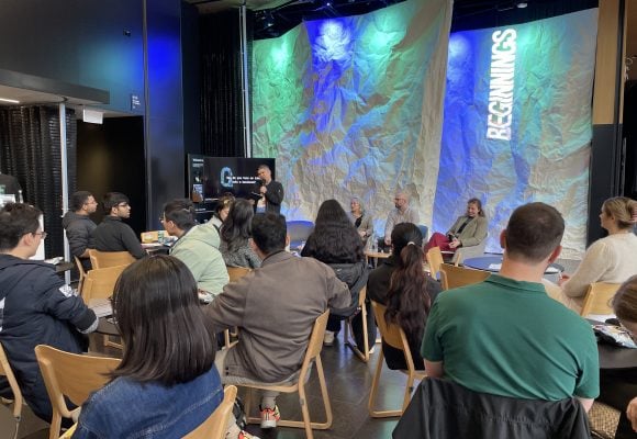 People sit at tables looking over to person speaking into a microphone