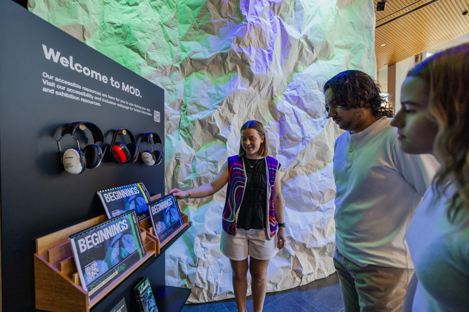 A group of three people look at a black stand which has three ear muffs on it and a document stand holding three guides.
