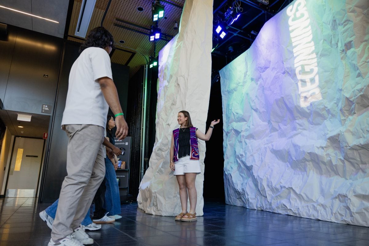 A tour guide stands a the entry to the BEGINNINGS exhibition which has two panels of crumple white walls. On one wall the word, 'BEGINNINGS' shines. A group of people to the left of the frame are walking toward the tour guide.