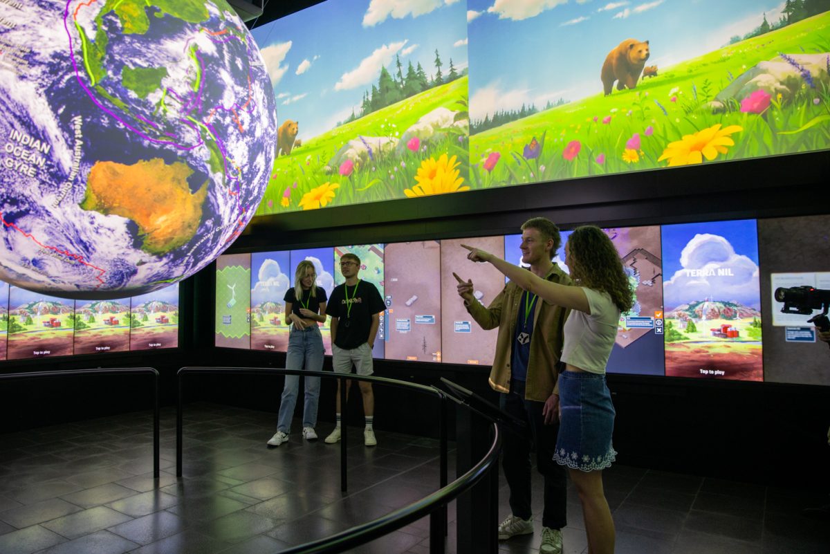 Four young people in a room of digital screens and projections, with two pointing at a large sphere of the Earth