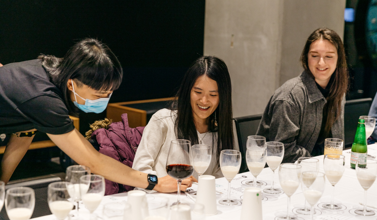 Two people sit at a table, surrounded by wine glasses filled with different plant-based milk. A third person leans over, pointing to something on a page. Everyone is smiling.