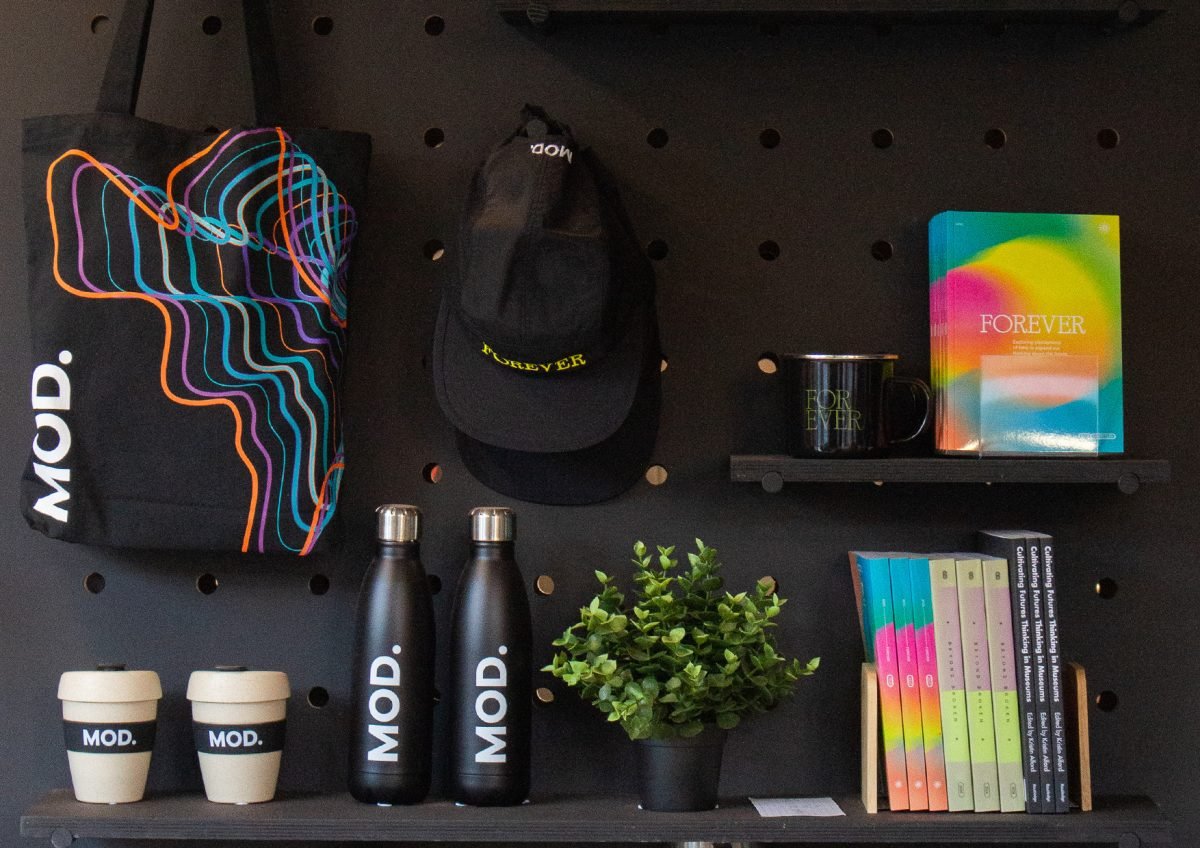 A black wall with a tote bag hanging down on the right, with light coloured keep-cups. In the centre is a black cap hanging, beneath it are black water bottles. To the left, is a blakc mug on a shelf with a FOREVER book next to it. Below that are more books with their spines showing and a small pot plant.