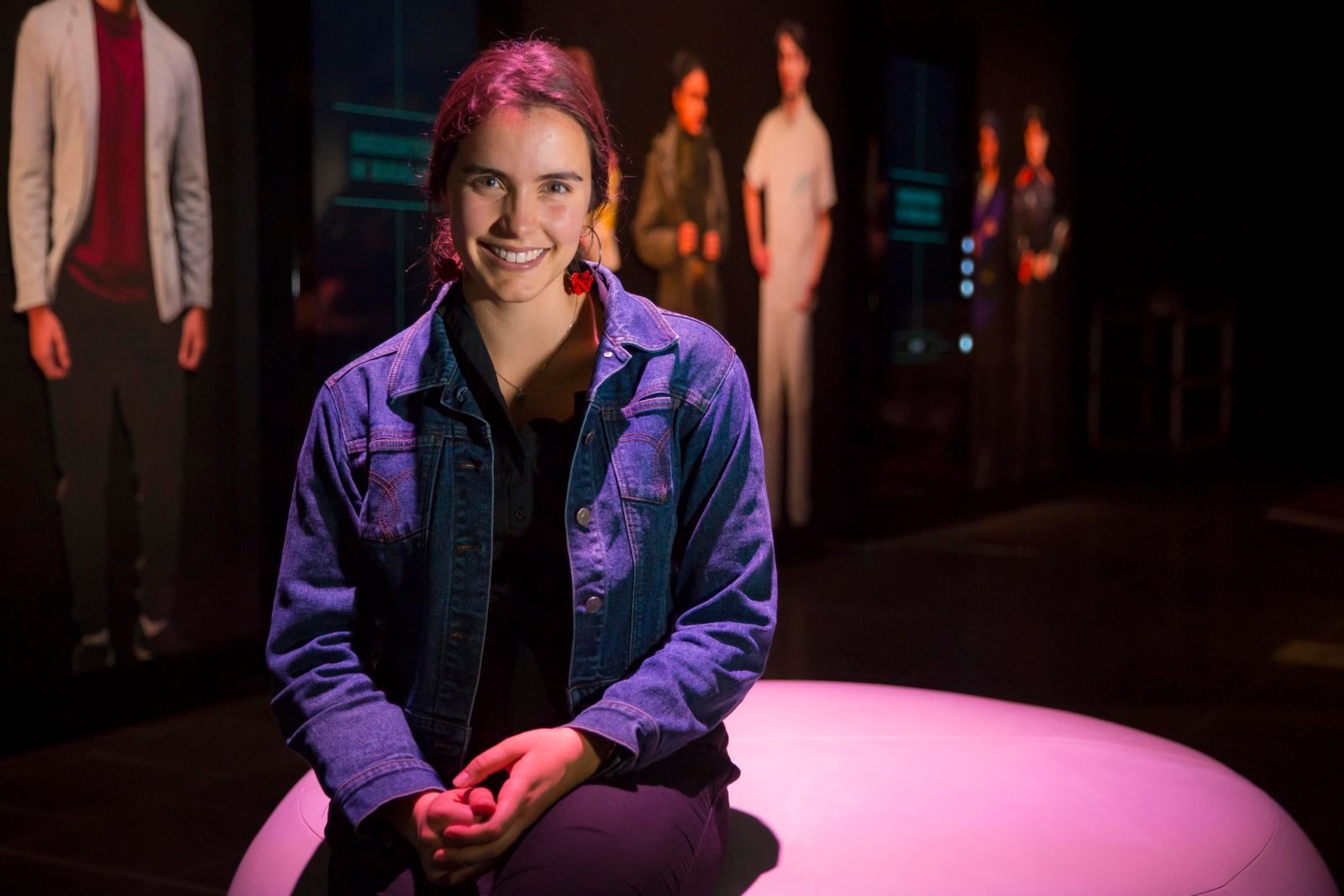 A young woman sits on a white, round bench and smiles at the camera. They are wearing a denim jacket and have their hair in a ponytail. The lighting is pink and washing over the photograph. 