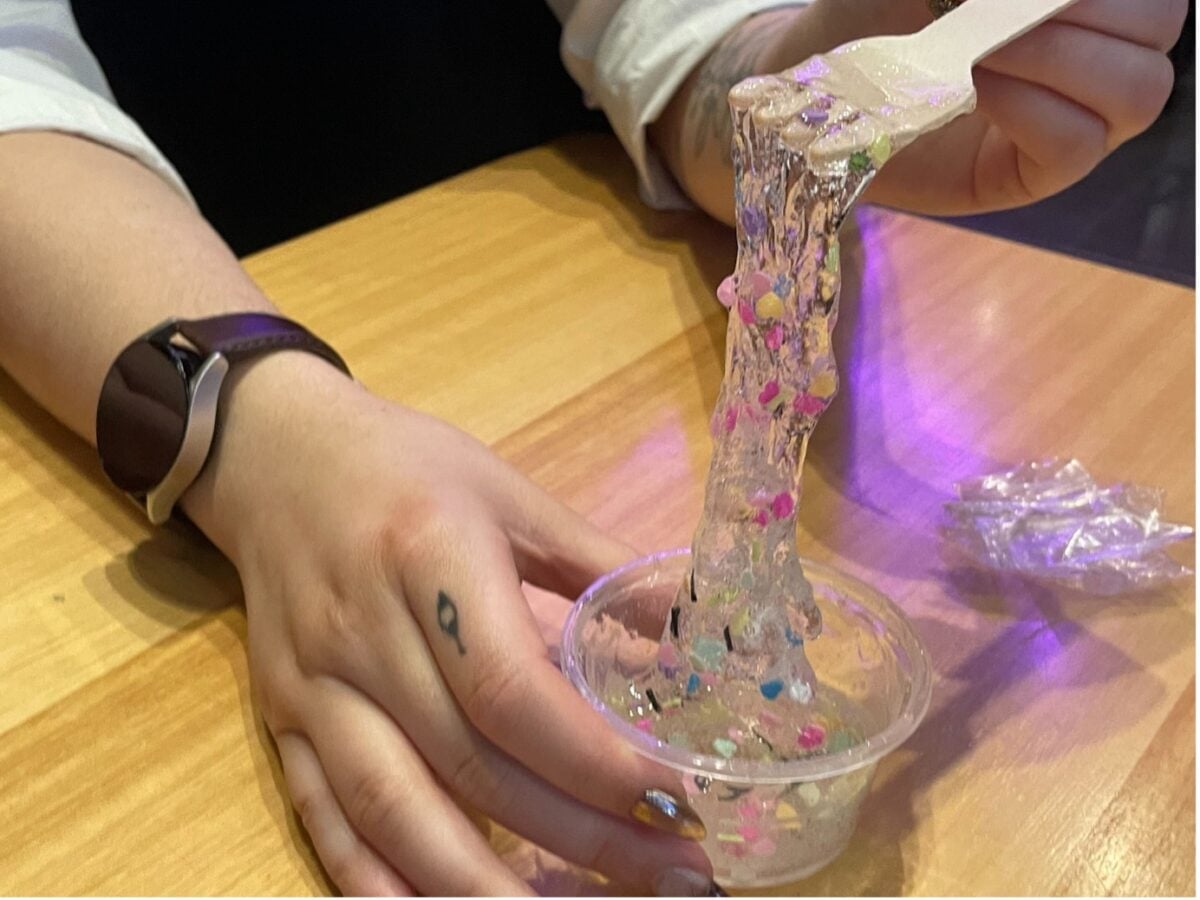 A hand holds a cup of slim with sparkles in it. The other hands use a fork to stretch out the slim.