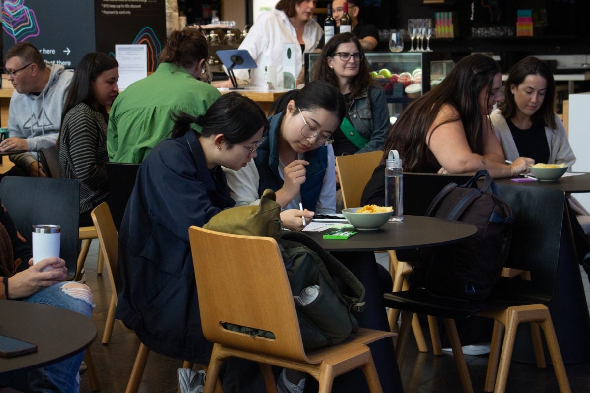 A crowd of people are participating in a quiz. in the middle are two people sitting a small round table. They are looking at a quiz page and working together to answer with a pencil poised above.