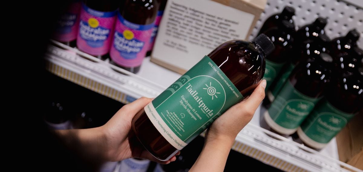 A dark brown shampoo bottle with a green label is held in the hands of a person. They are standing in front of white shelving with more of the same bottles and some with different labels stocked. There is a small cardboard box with writing on it sitting in the middle of the top shelf, this and the bottles are out of focus.