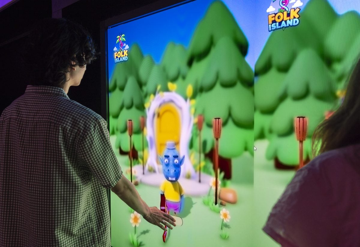 A person stands infront of a large screen with their hand outstretched to touch. The screen shows a small animated creature.