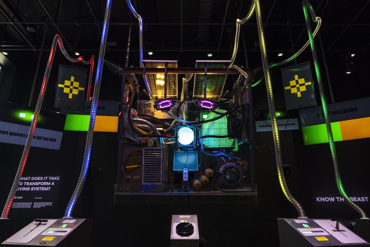 A large cubic creature made of lights, solar panels and industrial items looms at the camera with controls connecting to its body.