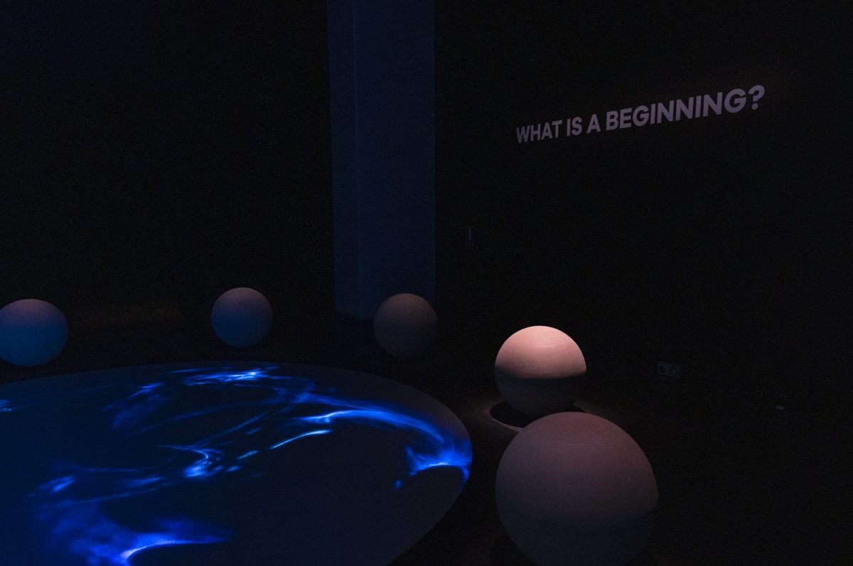 A dark room, with a floor projection of blue swirls. Stone spheres sit illuminated around the projection. Text on wall reads 'What is a beginning?'