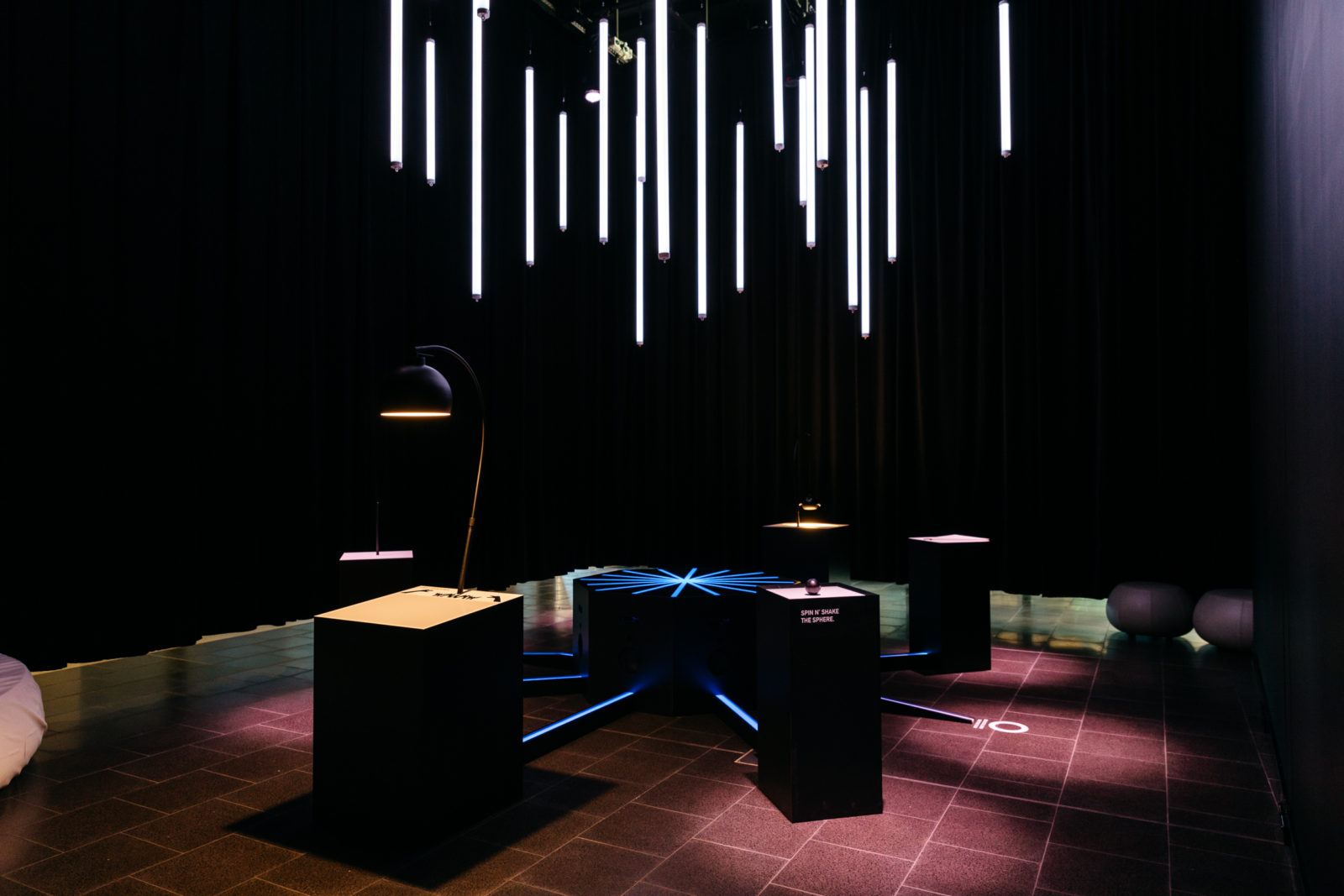 A dark room with long hanging light pendants in the centre. Around the edges are a number of plinths, each lit with a spotlight. There are strange instruments on each. 