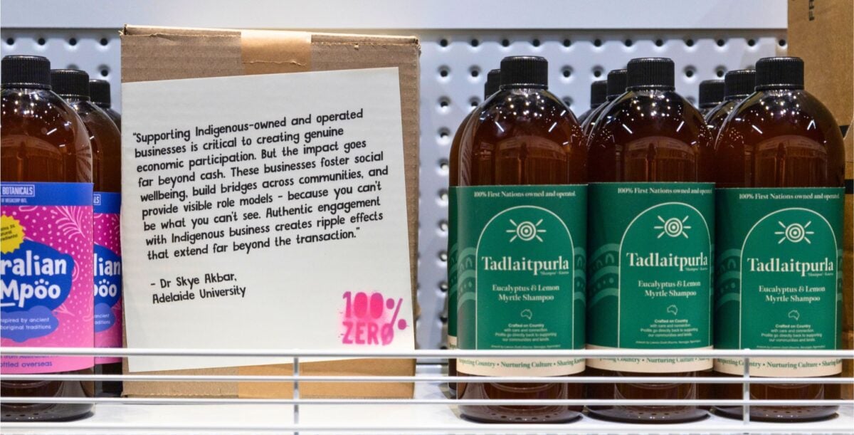 On white metal shelving there are two different shampoo brands, one with green labels and the other with pink and blue. Between the different brands there is a small cardboard box with a quote from a marketing researcher stuck to the front. There is a bright pink graffiti-looking tag that reads: "100% ZERO".