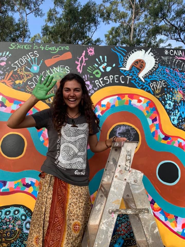 Tiahni smiling and holding up paint covered hand against backdrop of painted wall art