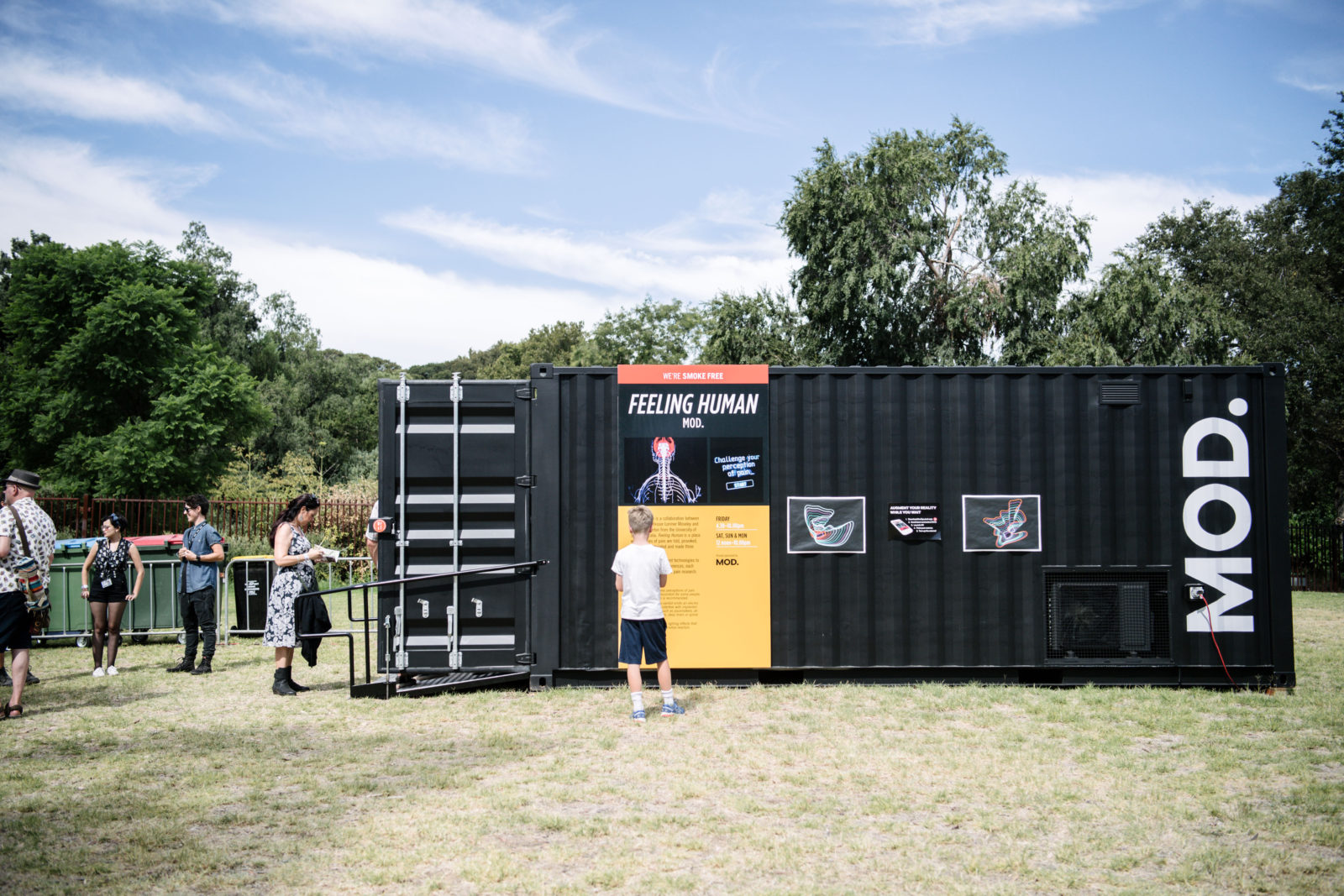 A black shipping container sits on grass. There is a queue of people waiting to get inside. The container is labelled MOD. and has information about the exhibit.