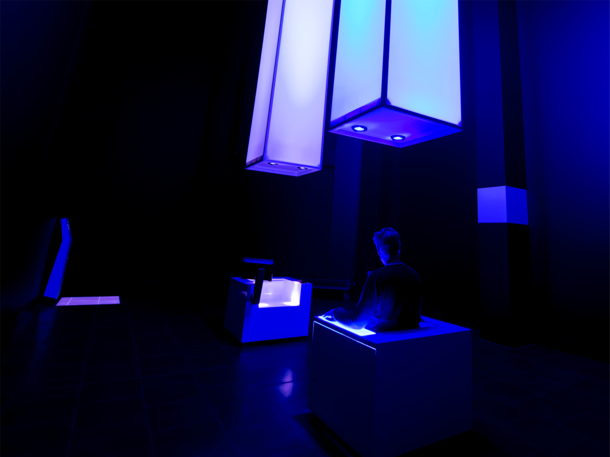 A dark room with two glowing columns hanging from the ceiling. A man is sitting in a chair with his back facing the camera.
